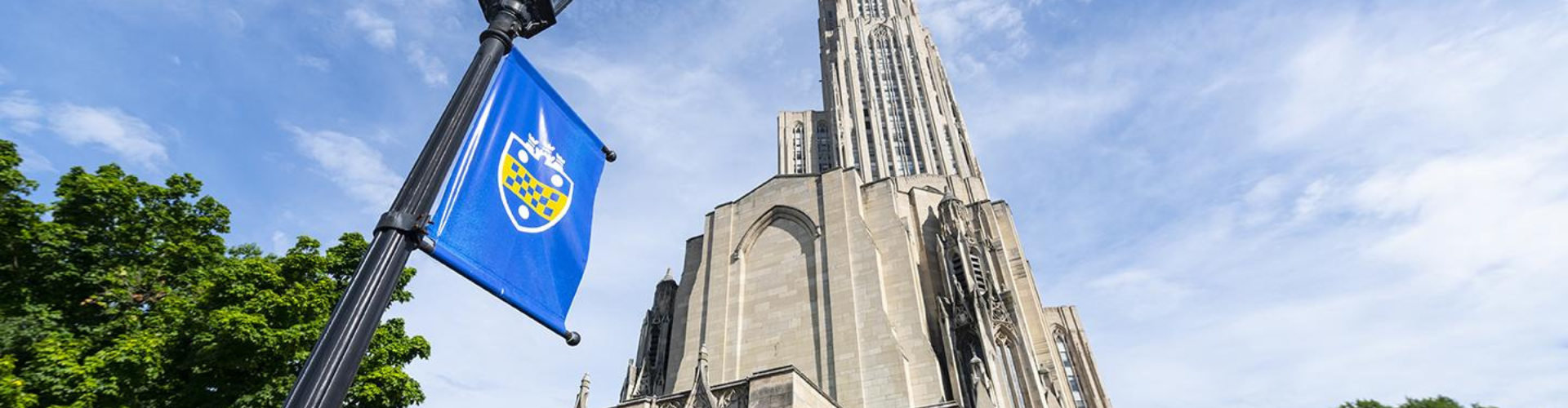 A Pitt flag foregrounds a low angle shot of the Cathedral of Learning against a blue sky.
