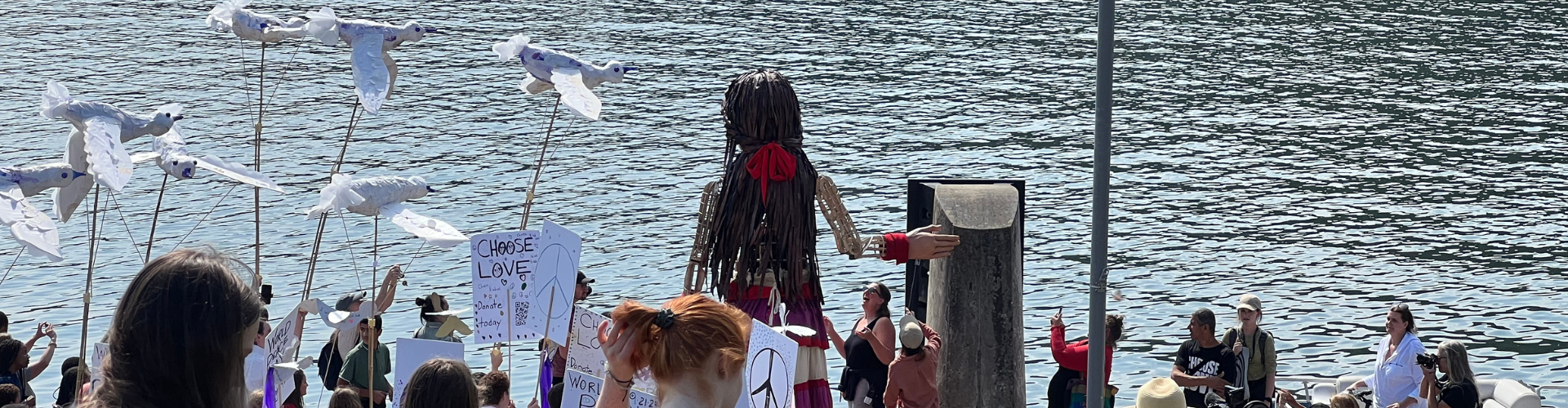 Little Amal, a large puppet, looks out across river, crowd of people behind her